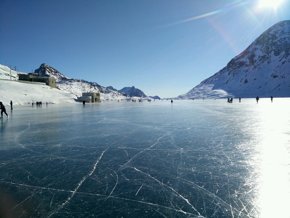 Lago Bianco frozen in winter at the Bernina Pass — people walking and skating on the ice, surrounded by snow-covered Alps